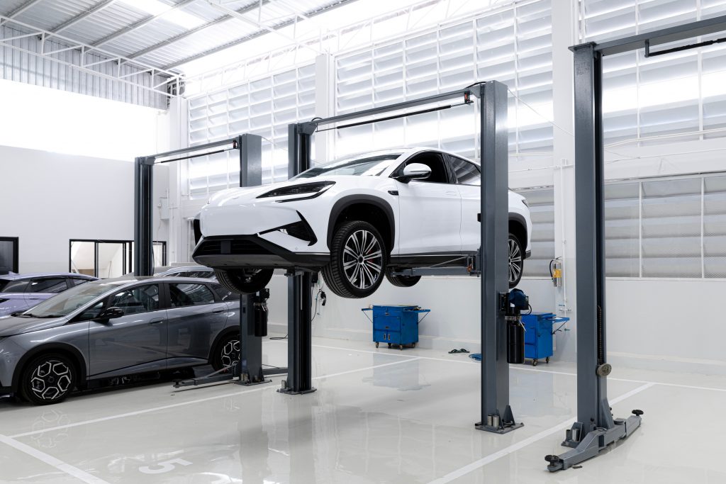 Electric SUV lifted on a hydraulic service bay inside a modern auto repair shop, with technicians’ equipment and additional vehicles in the background.