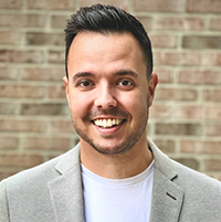 Professional headshot of a smiling man in a gray blazer and white shirt, photographed against a brick wall background.