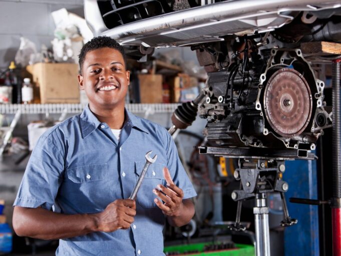 Smiling automotive technician holding a wrench in a repair shop, standing beside a vehicle lifted for engine or transmission service.