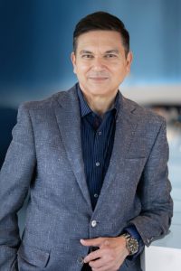 A professionally dressed man wearing a blazer and button-down shirt, standing indoors and smiling slightly at the camera.