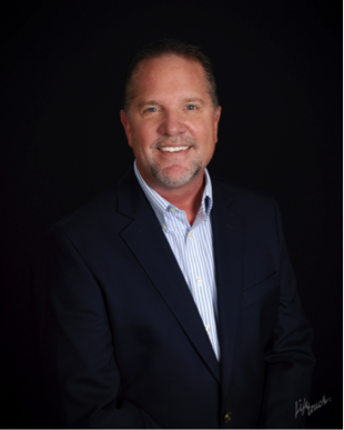 Professional studio headshot of a smiling, middle-aged man in a navy blazer and striped dress shirt against a black background.