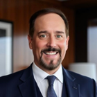 Professional headshot of a smiling man wearing a navy business suit and tie in a modern office setting with warm lighting.